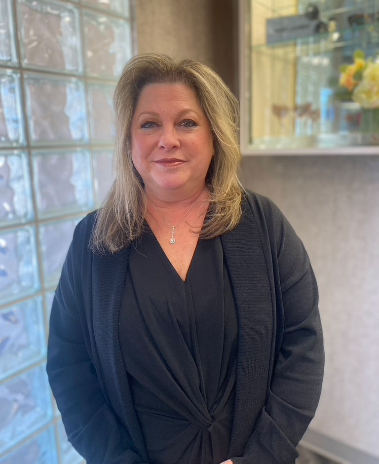 The image is a photograph of a woman standing in front of a glass window with a tiled interior, wearing a black blazer and posing for the camera.