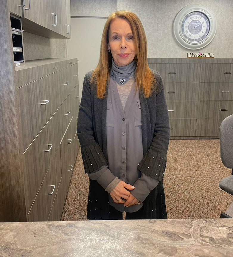 The image features a woman standing in an office environment, wearing a dark cardigan and a light-colored blouse. She has long brown hair and is looking towards the camera with a neutral expression.