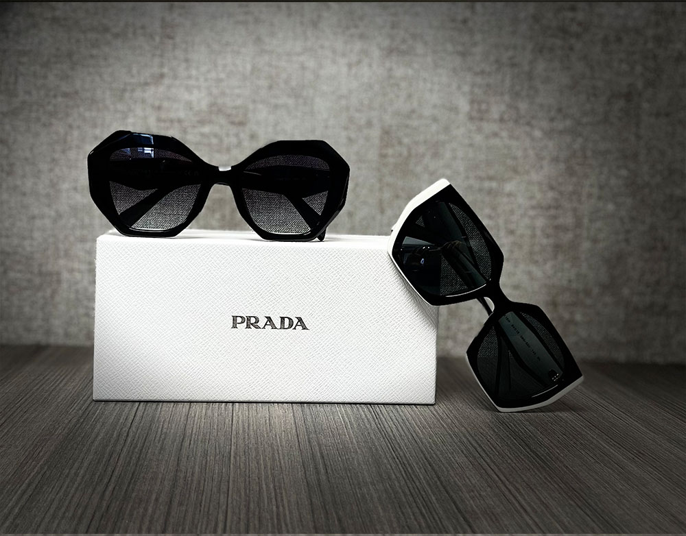 The image shows a box of Prada sunglasses, with two pairs displayed in front, against a neutral background.