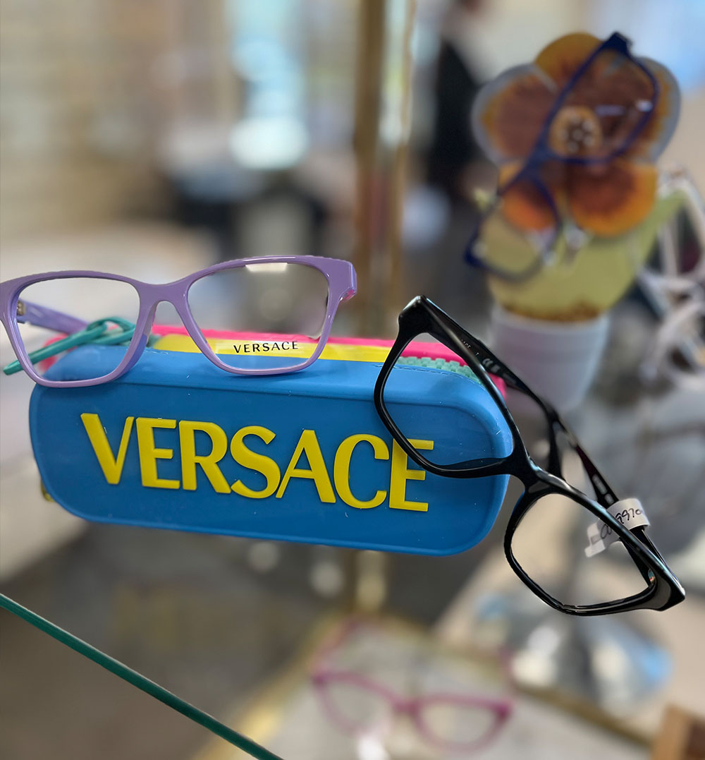 The image displays a collection of eyeglasses, including frames in various colors and styles, with one pair prominently featuring the word 'VERSACE' on its case.