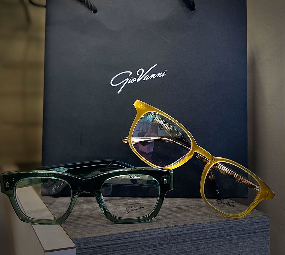 A display of two pairs of eyeglasses, one yellow and the other green, with a sign in the background that reads 'GIOVANNI'.