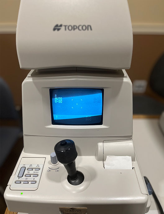 The image shows a piece of medical equipment, specifically an ophthalmic device, with a screen displaying text and icons. It is situated on a table indoors.