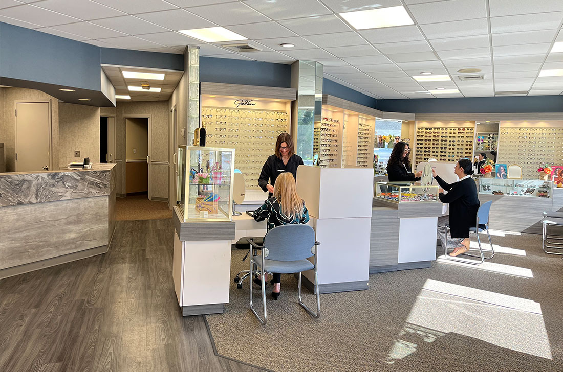 The image depicts an interior view of an optician s office with a woman standing behind the counter and multiple chairs arranged in front of a desk.
