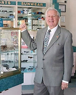 The image shows an elderly man in a suit standing behind a display case containing various items, likely within a store or exhibition space.