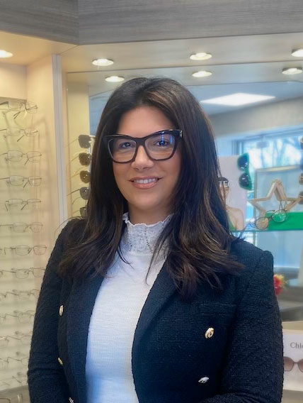 The image shows a woman standing in front of an optical store display, wearing glasses and a dark blazer with a lighter shirt underneath.