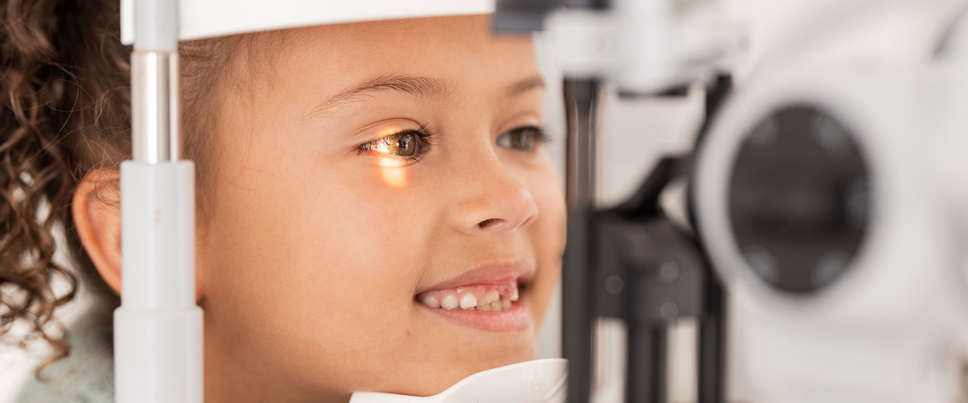 A young child, possibly a girl, is seated in front of an eye exam machine, looking attentively through the device with interest and curiosity.
