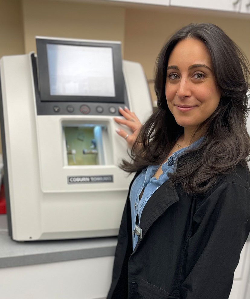 The image shows a woman standing next to a piece of equipment, likely in a laboratory or scientific setting.