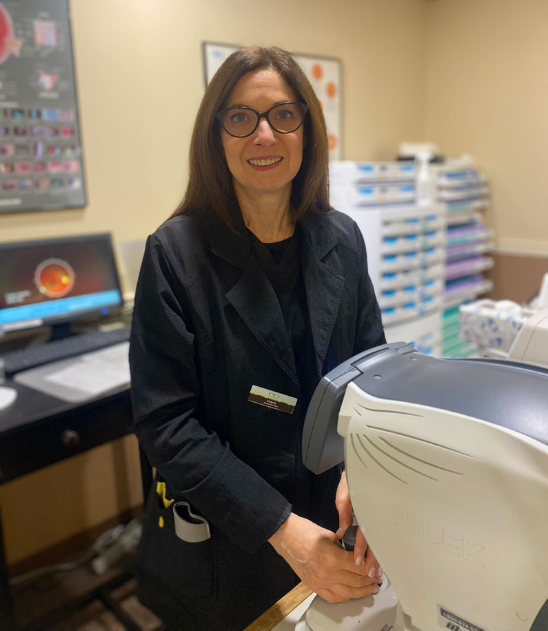 In the image, there is a woman standing at a counter in what appears to be an office or medical setting. She has dark hair and is wearing glasses, a black jacket, and a name tag. The woman is smiling slightly and looking towards the camera. Behind her, there are various items on the counter, including what looks like a computer monitor and some paperwork. To her right, there is a machine with a screen displaying medical imaging.