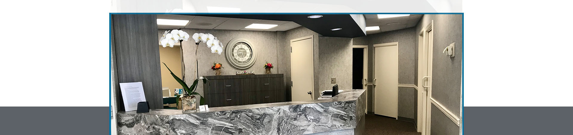 The image shows an interior space, likely a lobby or waiting area, with a modern design featuring a large countertop with a built-in desk and chair arrangement. There is a marble-like patterned floor, white walls, and a decorative ceiling light fixture. A red fire extinguisher is mounted on the wall next to the desk. The space is well-lit and appears clean and organized.